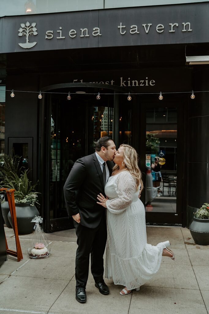 Bride and Groom kiss in front of Siena Tavern restaurant with string patio lights above them. 