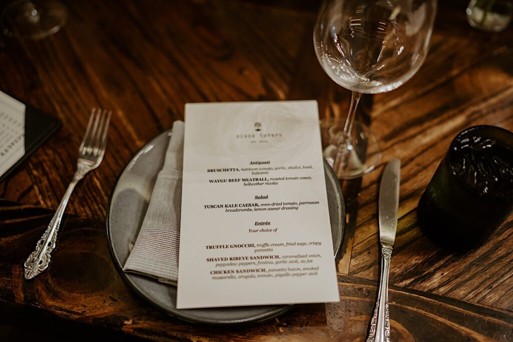 On a wooden table sits a plate with a Siena Tavern menu and utensils sitting on either side. 