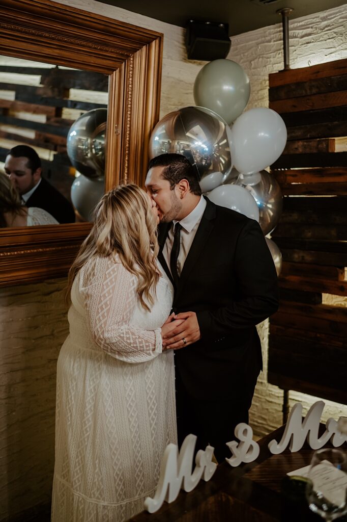 Bride and Groom kiss in-front of silver and white balloons set up in a private dining room for their intimate brunch reception.