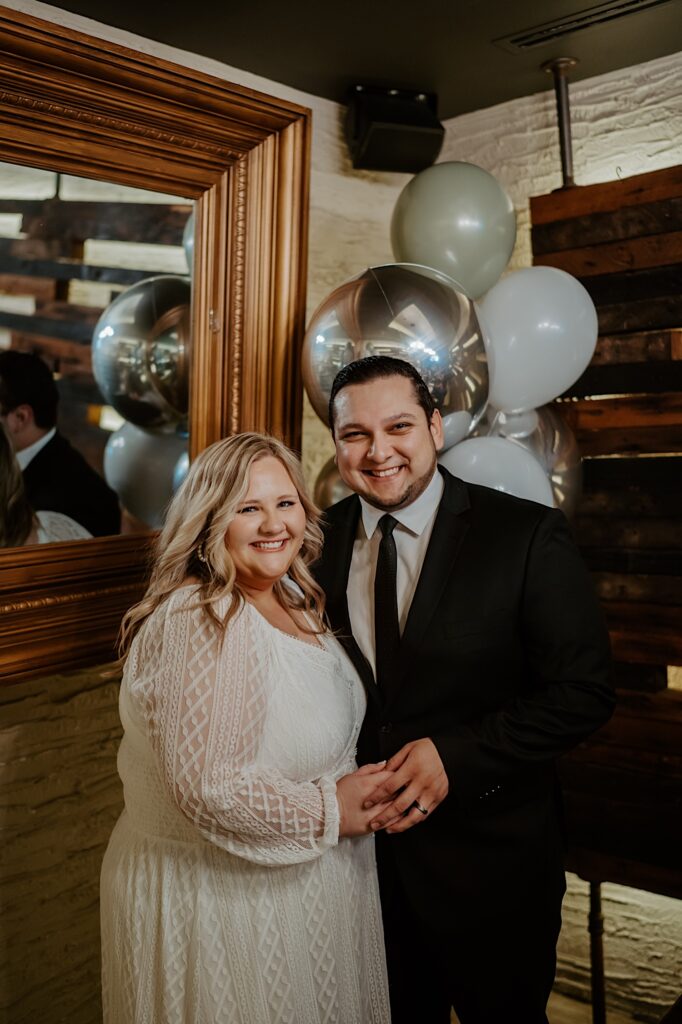 Bride and Groom smile in-front of silver and white balloons set up in a private dining room for their intimate brunch reception.