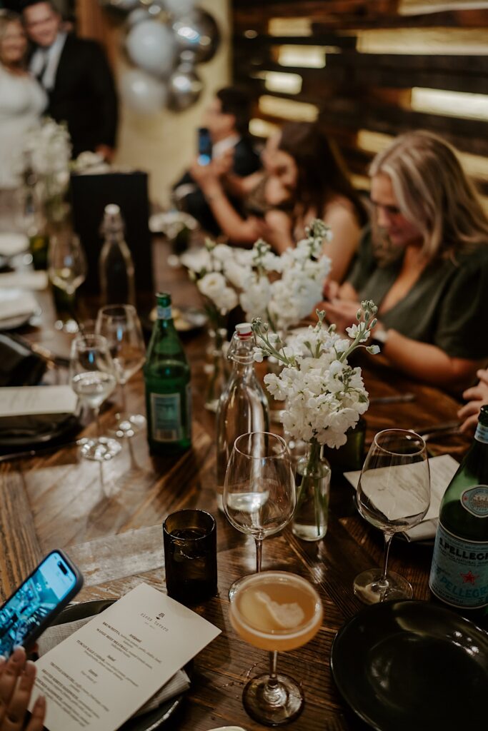 Guests sit around a wooden table with cocktails and vases of white flowers during intimate elopement reception at Siena Tavern. 