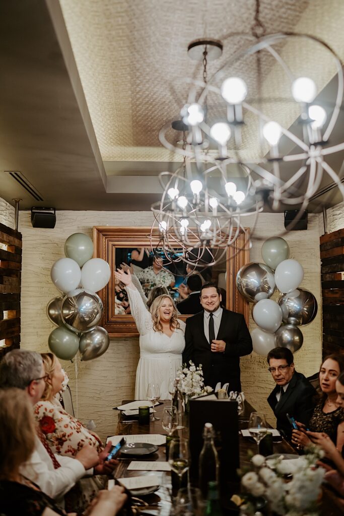 Bride holds her arm up in celebration as she stands next to the groom and her guests sit at a wooden table in front of them. 