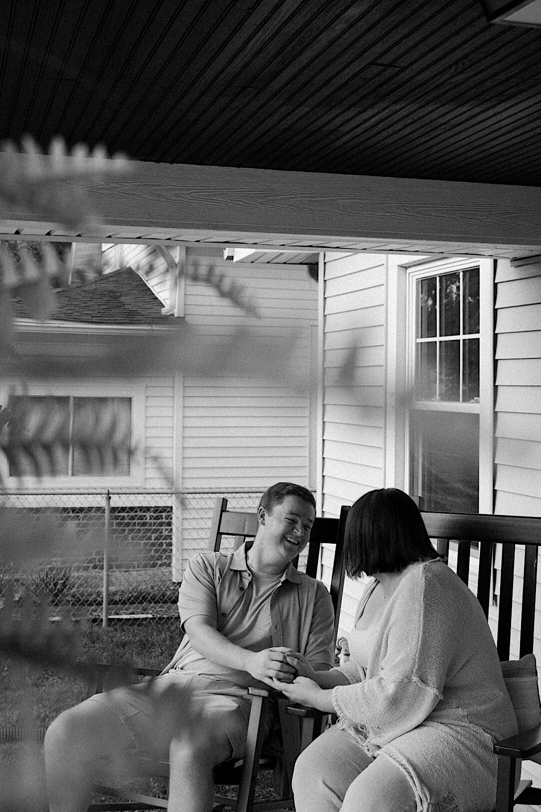 Couple holds hands as they sit on rocking chairs on their porch and laugh while looking at each other candidly. 