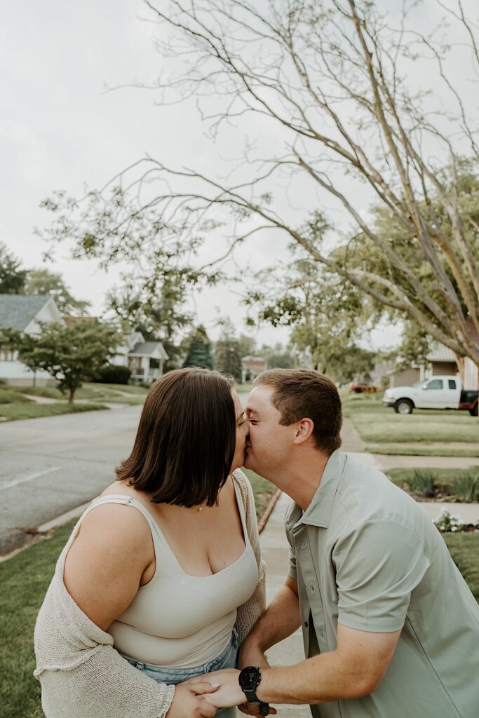 Couple kisses while walking down the sidewalk in their neighborhood for cozy portraits by Indiana engagement photographer. 