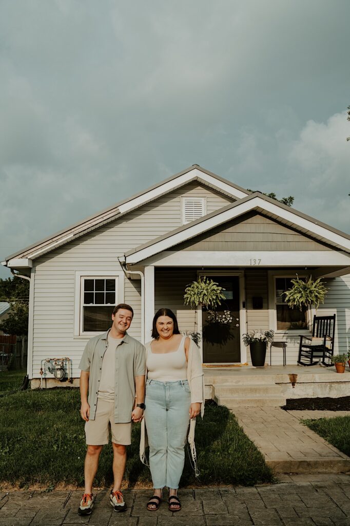 Couple poses in front of their house and smiles at Indiana engagement photographer in Lafayette, Indiana. 