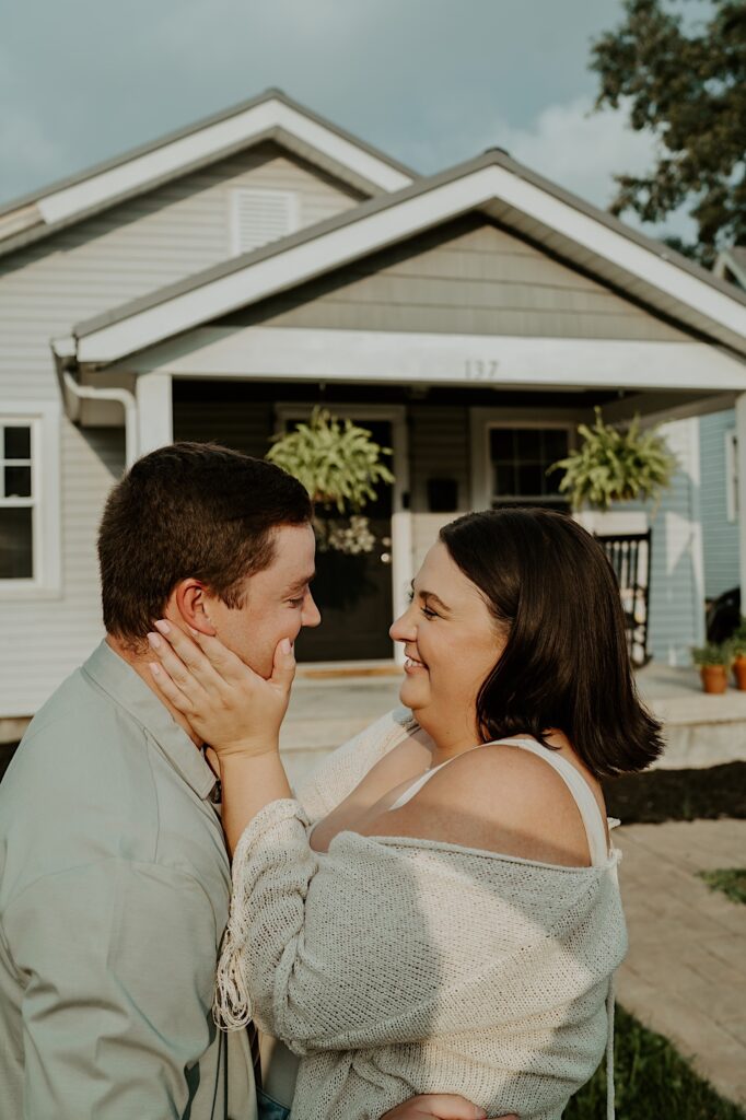 Woman holds her husbands face as they stand in front of their house in Indiana for cozy date night portraits. 