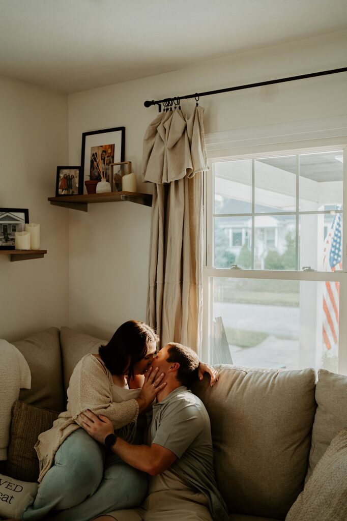 Couple sits on their beige couch in their living room and kiss during date night portraits in Indiana.