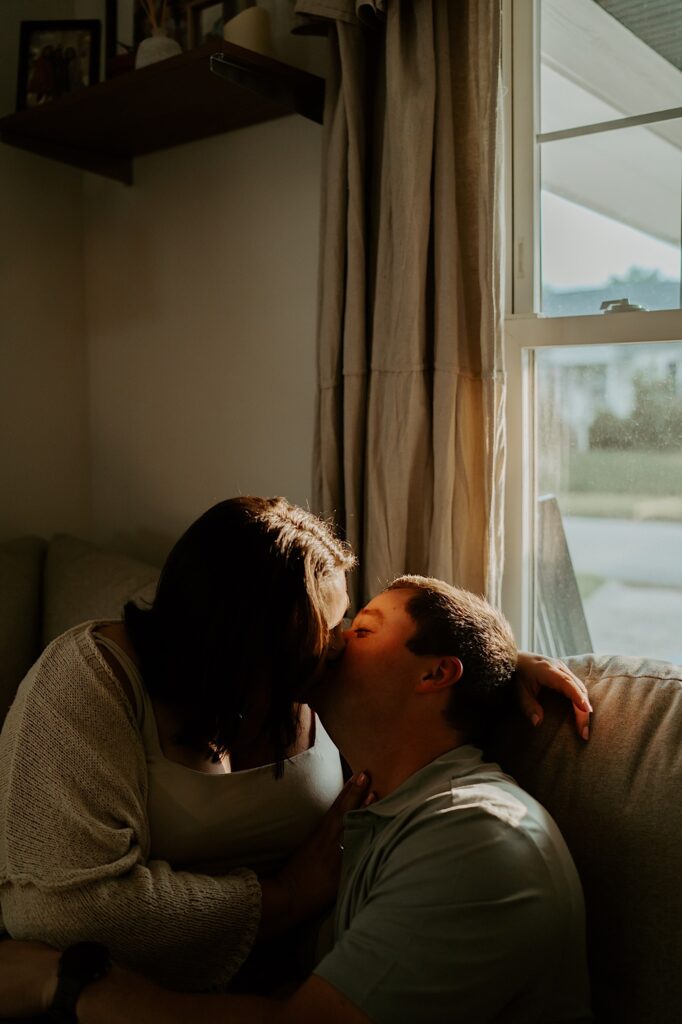 Couple sits on their beige couch in their living room and kiss during cozy date night portraits in Indiana.