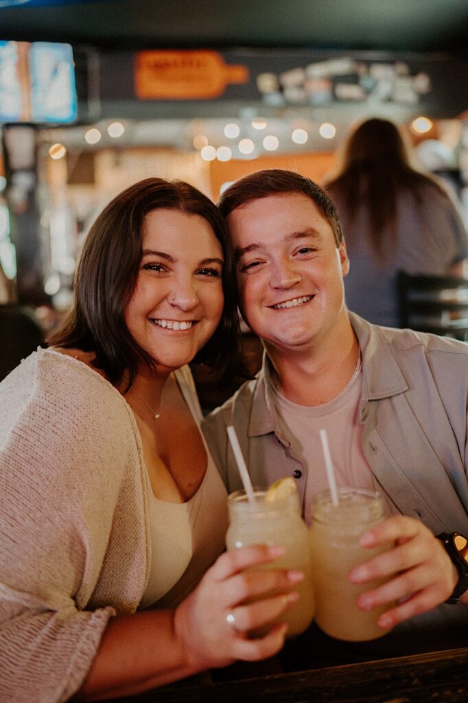 Couple holds margaritas in mason jars and smile at Indiana couple photographer during their date night at Sixth St Dive Bar & Grill in Lafayette, Indiana.