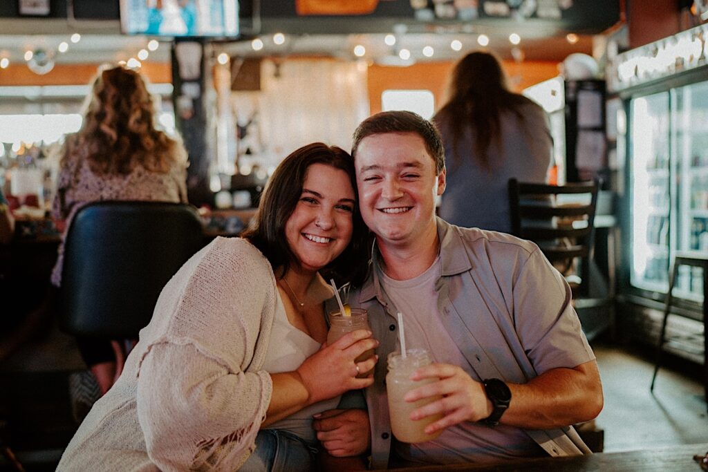 Couple holds margaritas in mason jars and smile at Indiana couple photographer during their date night at Sixth St Dive Bar & Grill in Lafayette, Indiana.