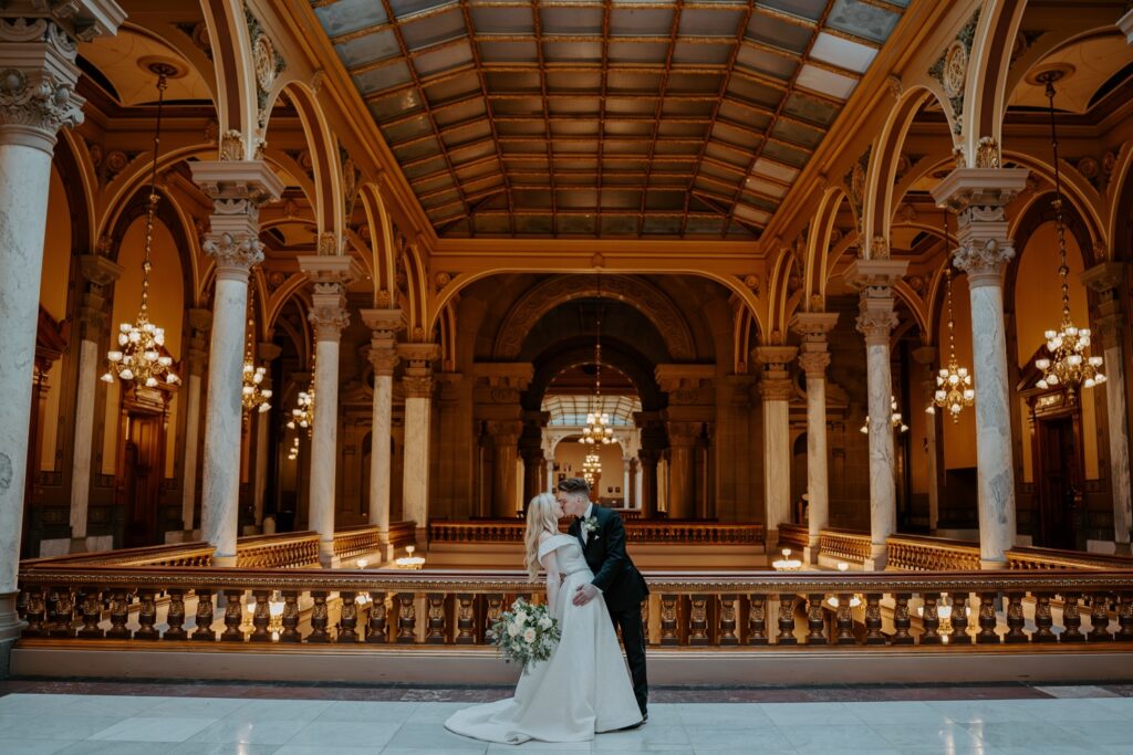 Bride in princess style dress kisses groom while holding bouquet of white roses and greenery in the upper level of the Indiana State House. 