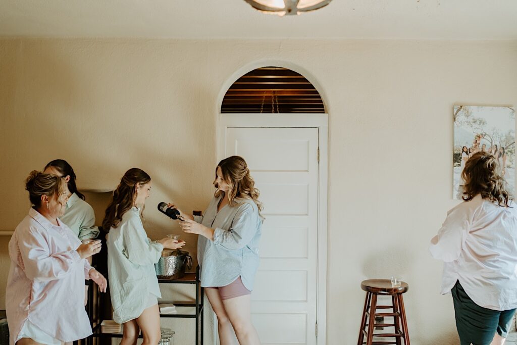 A bride gets ready with her bridesmaids at Mt Woodson Castle in Ramona California.  They're wearing pastel colored button up shirts and pouring champagne to celebrate.