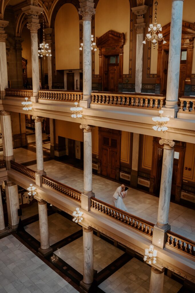 A bride and groom kiss on the second level of the Indiana State house.  The bride wears a short veil with her hair in a high bun. 