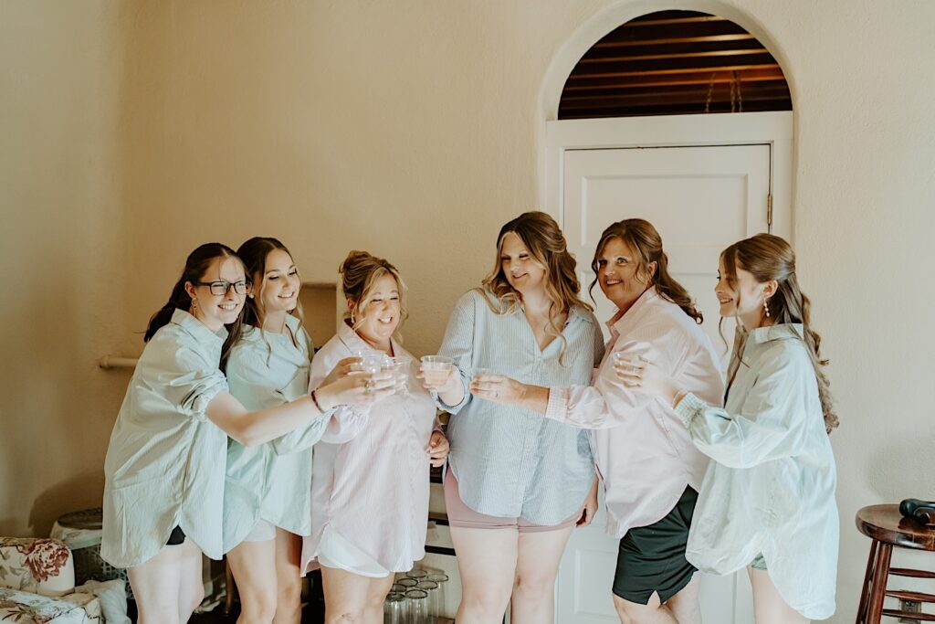 A bride and her bridesmaids toast to her wedding day dressed in matching oversized button up shirts at their California wedding venue. 