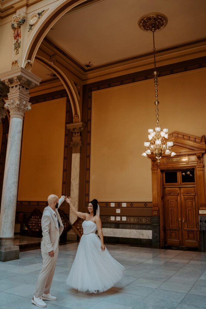 A bride and groom dance at the Indiana State House after their elopement.  The groom wears a khaki suit with sneakers and the bride wears a two piece wedding dress with a heart shaped corset top and a full tulle skirt.