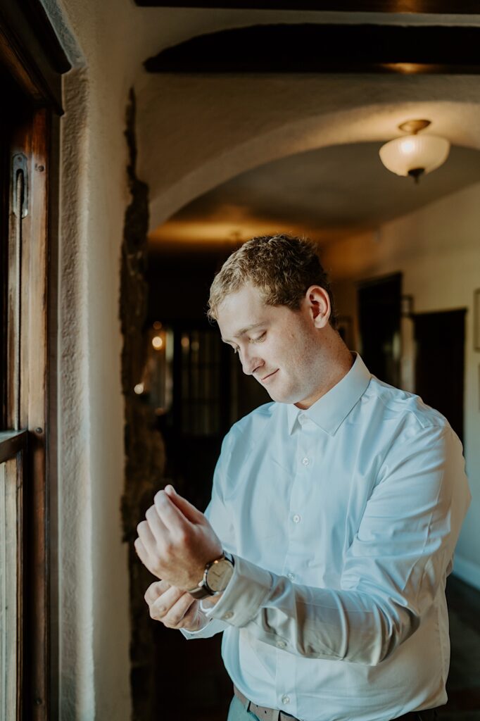 The groom fastens his watch standing near a window at Mt Woodson Castle in Ramona California