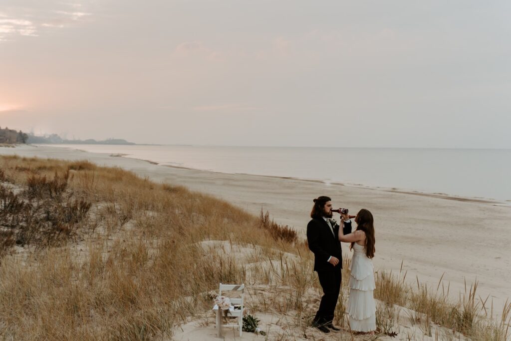 A bride and groom toast to their marriage with beers overlooking the Indiana Dunes on the coast of Lake Michigan.  The groom wears a black suit with a white button up and a boutonniere and the bride wears a dress with layered pleated fabric and her hair is in loose curls.