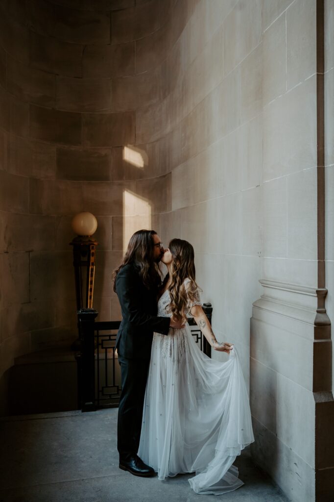 A bride in a dramatic wedding dress dress holds it out showcasing the unique texture and kisses her husband in all black in a hallway in the Indiana State house. 