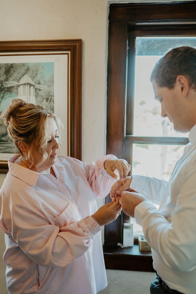 The mother of the groom helps him put on his cuff links the morning of his wedding day.  The mother of the groom is wearing an oversized getting ready button up shirt.  They're in the getting ready space at  Mt. Woodson Castle California.