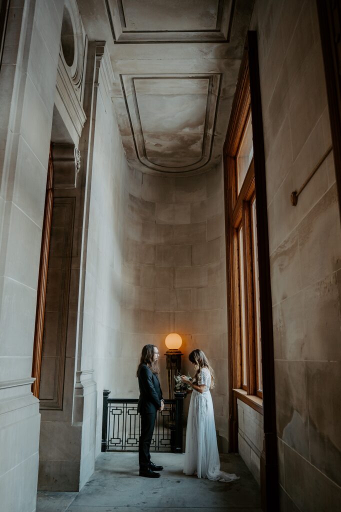 A bride in a dramatic wedding dress dress holds it out showcasing the unique texture and shares vows with her husband in all black in a hallway in the Indiana State house. 