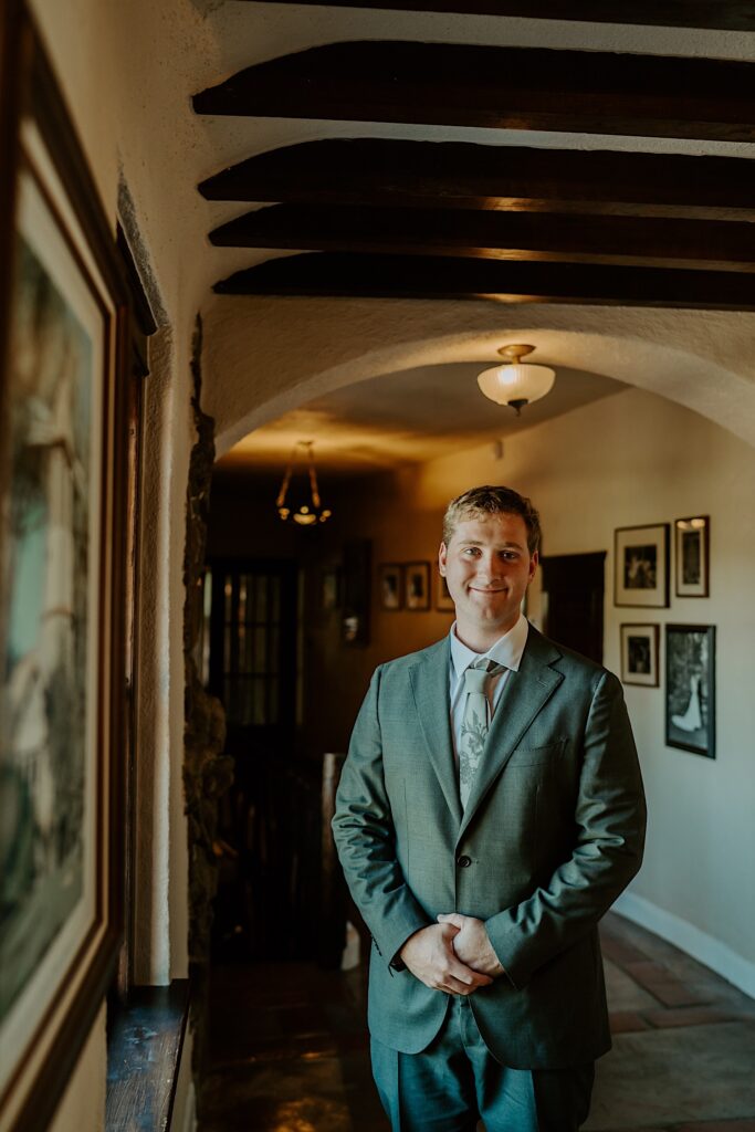 A groom stands in the window after getting ready at his California wedding venue.  He wears a charcoal grey suit with a green patterned tie.