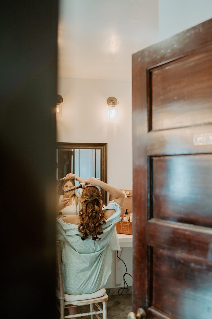 A bride gets ready in the getting ready suite at Mt Woodson Castle in California.  She's wearing a light blue button up and curling her hair. 