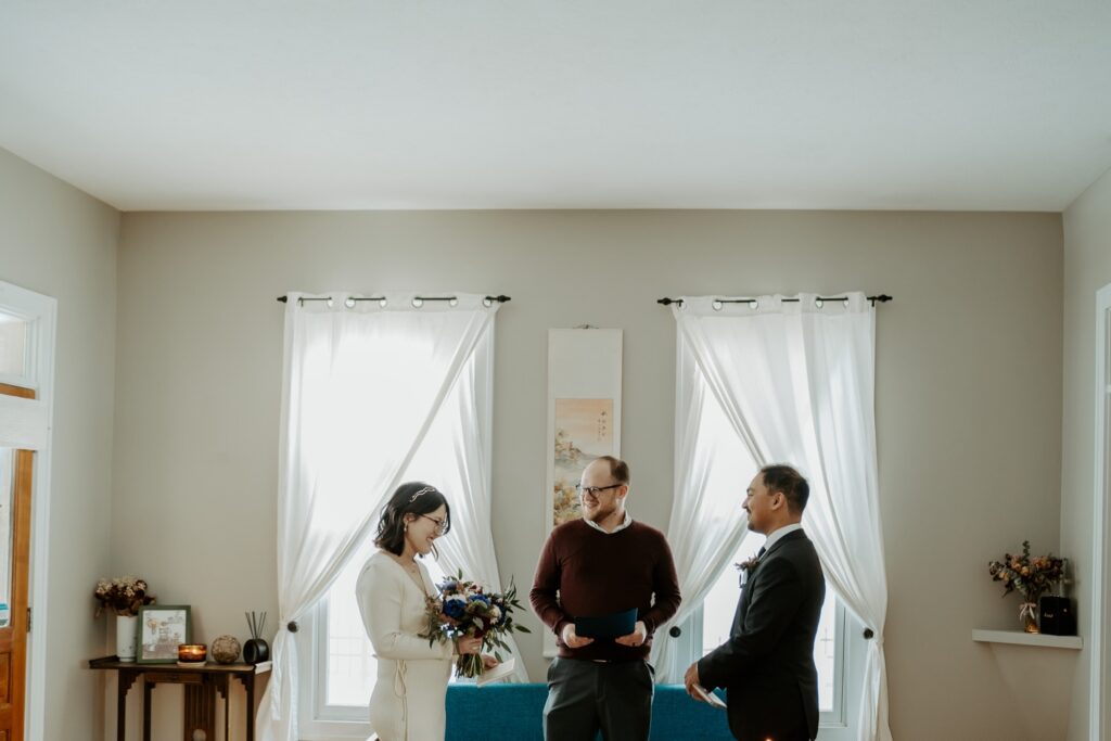 A couple elopes during an intimate ceremony in their Indianapolis home on a winters day.  The bride wears a simple knit white dress and the groom wears black tie attire.  They are framed by two windows in their Indianapolis bungalow. 