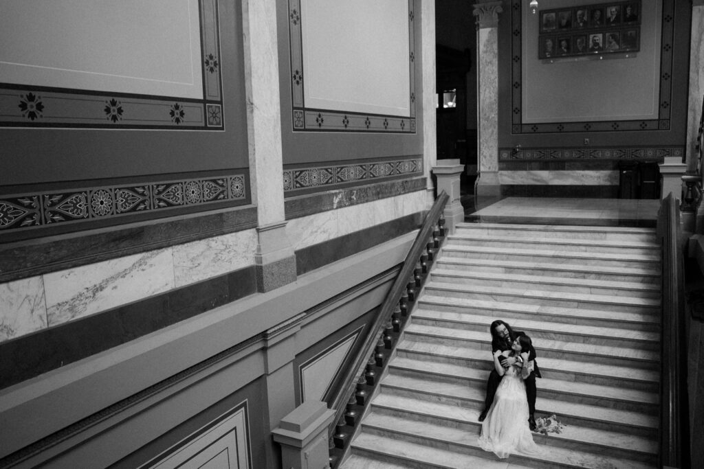 A couple sits on the marble steps of the Indiana State house.  The brides bouquet sits on the steps next to them and her dress falls down a step below them.  