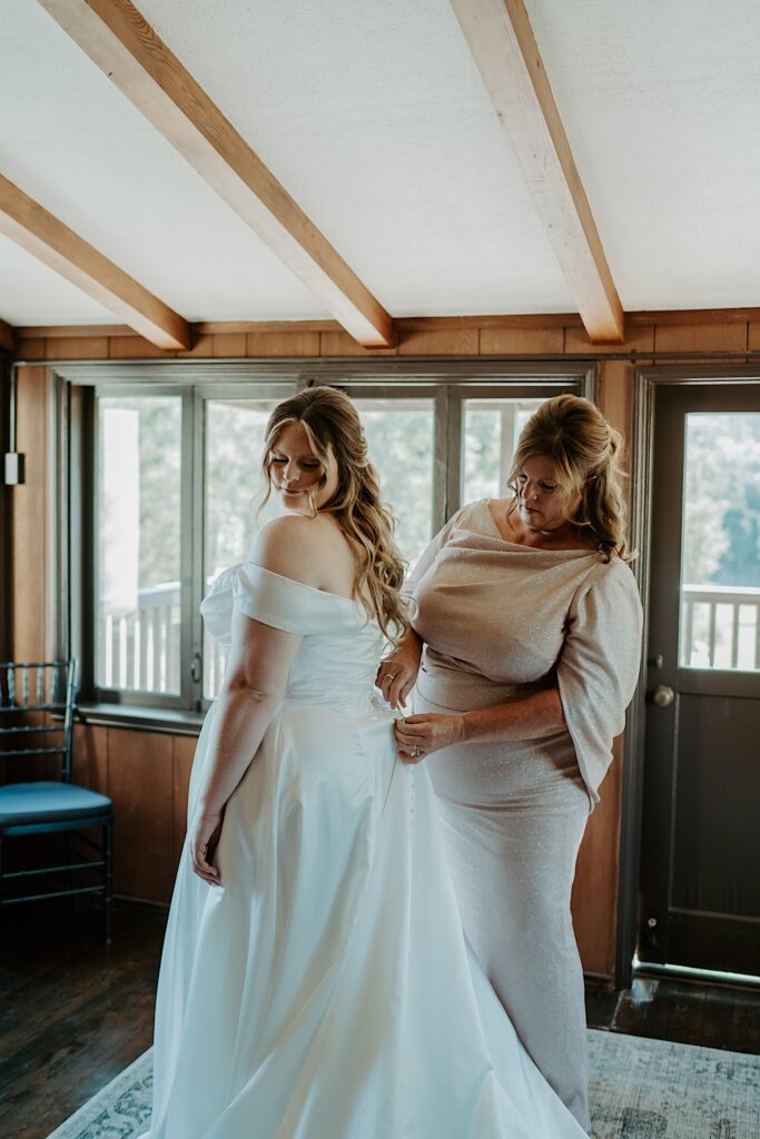 A bride has her mom put on her wedding dress in the getting ready suite at Mt Woodson Castle.  The mother of the bride is wearing a champagne colored sparkling dress.