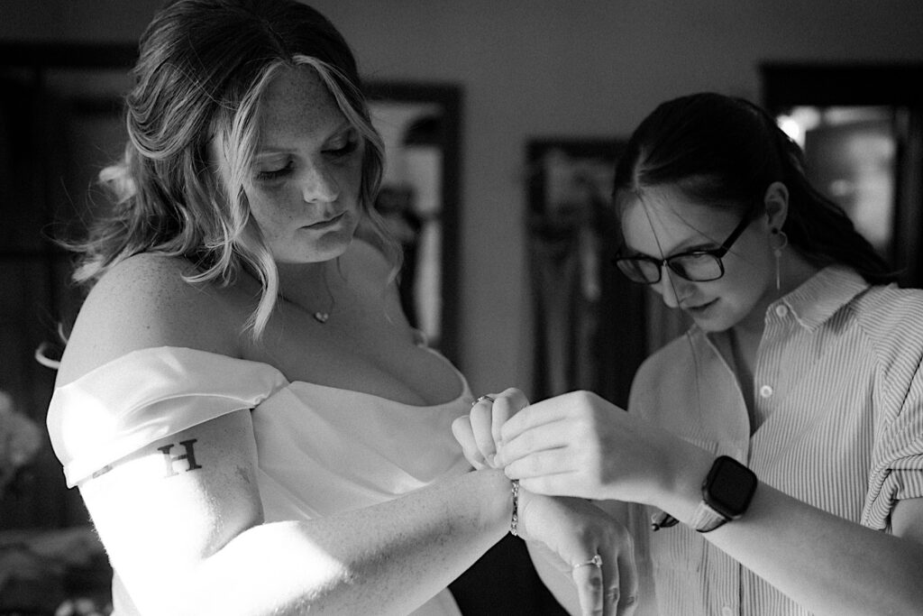 A black and white image of bride has a bridesmaid help her put on her wedding jewelry.  The bridesmaid is wearing an oversized pinstriped button up top and the bride is wearing a strapless dress with draping sleeves.