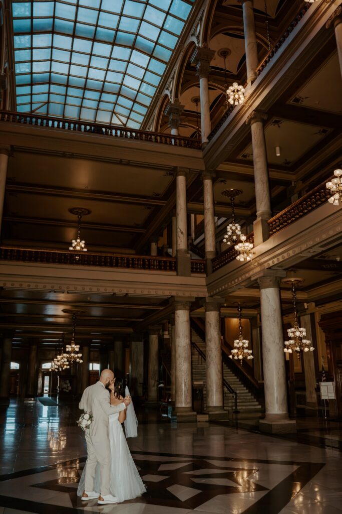 A bride and groom kiss and dance in the center of the Indiana State Houses' great hall. The bride wears a two piece dress with a veil that hits at her waist.  The groom wears a beige wedding suit. 