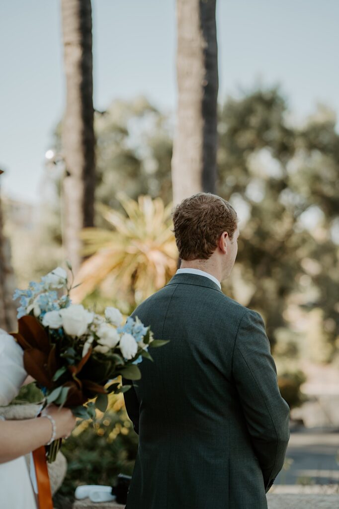 A groom waits as his bride approaches him before their first look on a sunny day in California.  The groom is wearing a charcoal grey suit and the bride is carrying a bouquet with blue and white flowers.