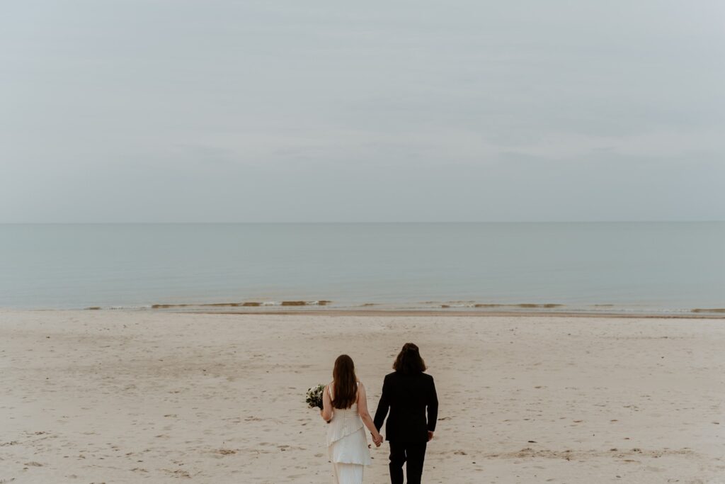 A bride and groom hold hands facing  Lake Michigan at the Indiana Dunes. The bride wears a pleated dress with multiple layers.