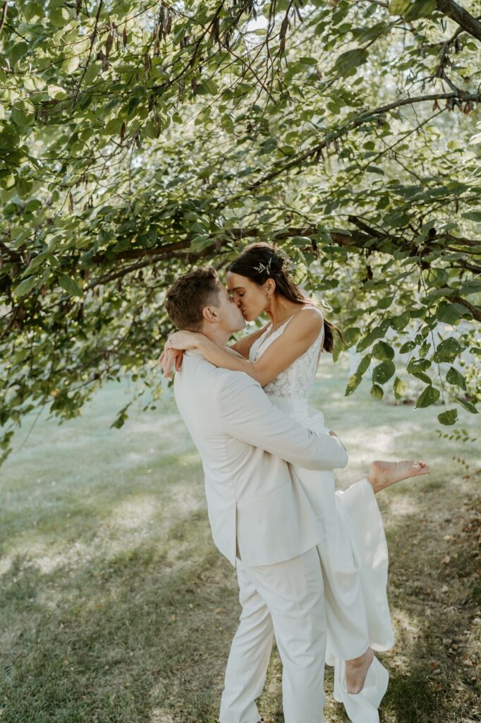 A bride and groom kiss after their backyard elopement ceremony in Indiana.  The bride has flowers in her hair and is barefoot.  The groom is wearing a beige linen suit. 