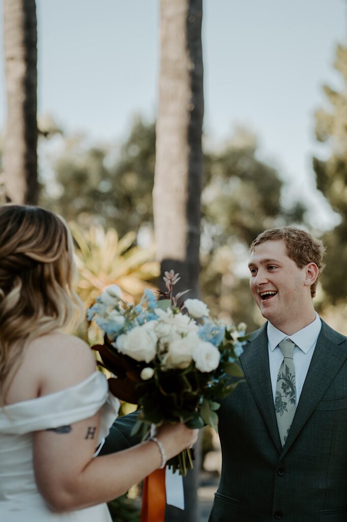 A groom reacts to seeing his bride for the first time on their wedding day as she approaches him for their first look on a sunny day in California.  The groom is wearing a charcoal grey suit and the bride is carrying a bouquet with blue and white flowers.