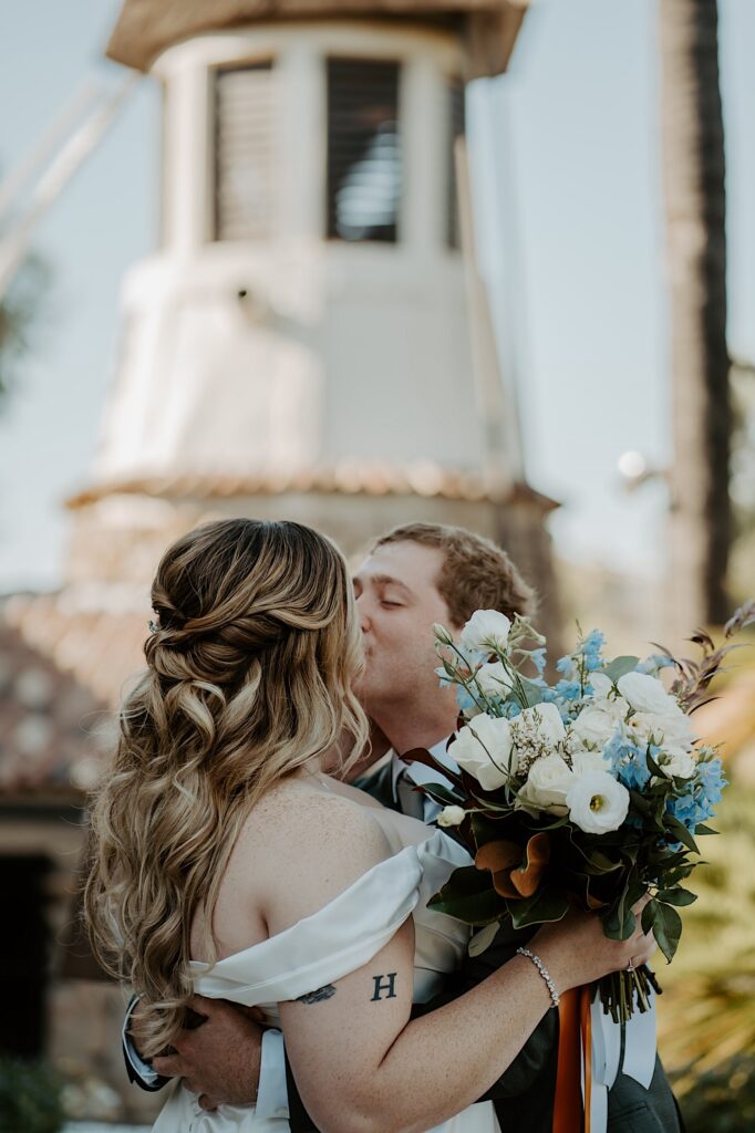 A bride and groom kiss during their first look on their wedding day during the summer in California.  The bride is wearing a loose curled half up half down hairstyle and holding her white and blue bouquet.