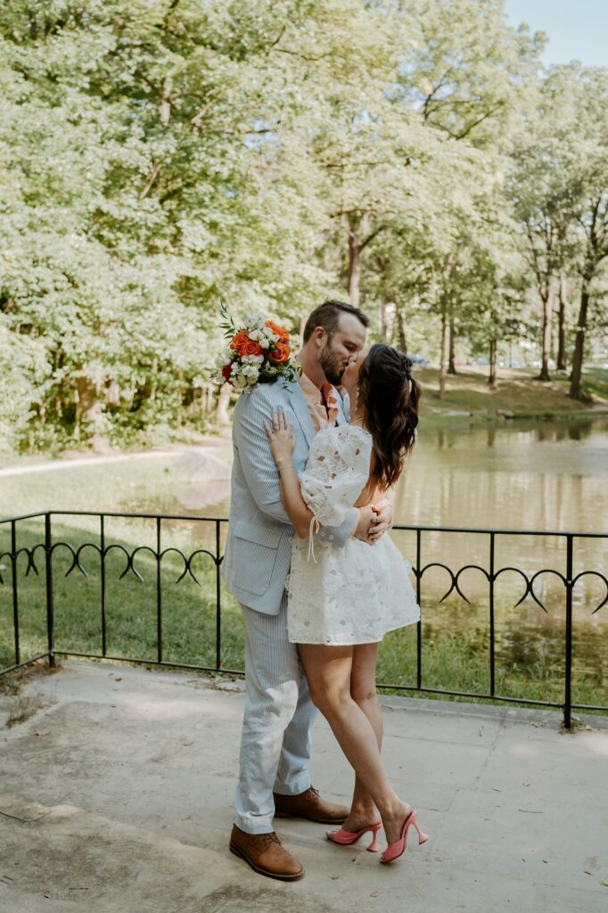 A bride and groom elope with a witness at Holcomb Gardens in Indianapolis.  The bride kisses her groom, she's wearing a springy floral lace dress with peach heals, while her groom wears a baby blue linen suit with a peach shirt.  She holds a bouquet of white and orange flowers.