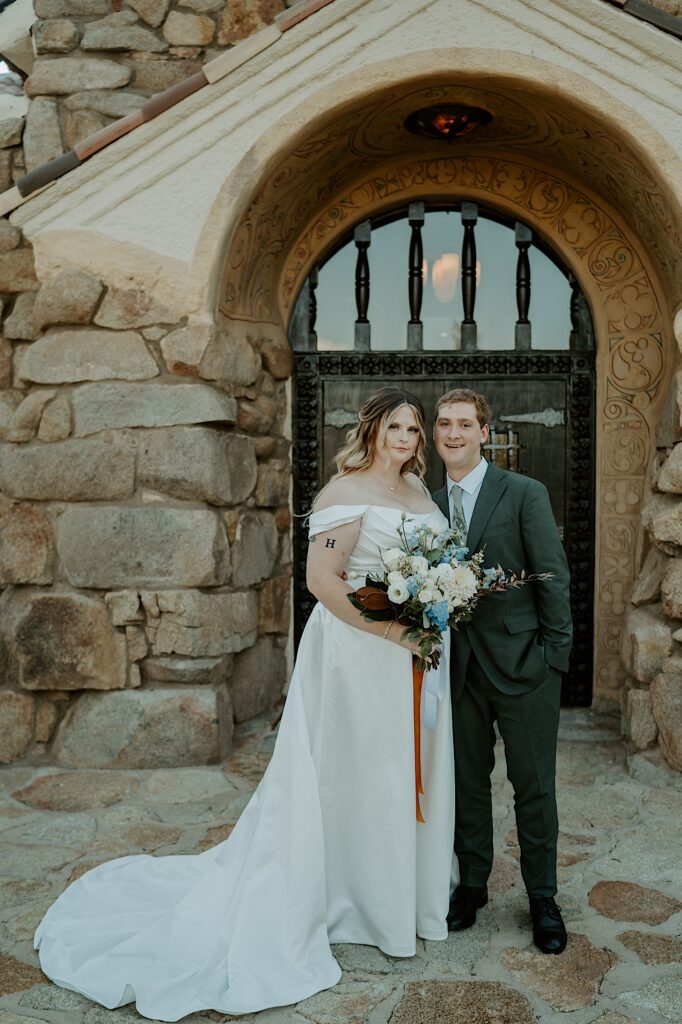 A bride and groom stand in front of an ornate door framed with large stones during their destination wedding at Mt Woodson Castle in California.  The bride is wearing a romantic dress with draping sleeves and a long train and carrying a bouquet with blue and white flowers.