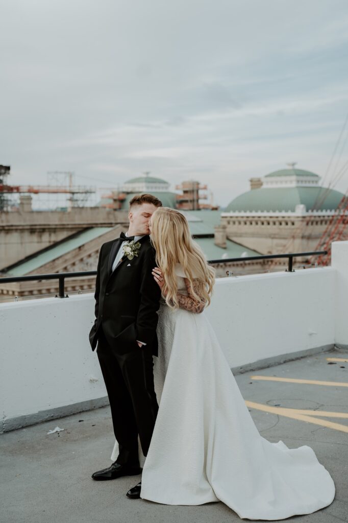 A bride and groom kiss on a rooftop parking lot in the center of Indianapolis. The bride wears a princess style dress with draping sleeves that accentuate her full sleeve of tattoos.  She wears her hair down in loose curls for her wedding day. 