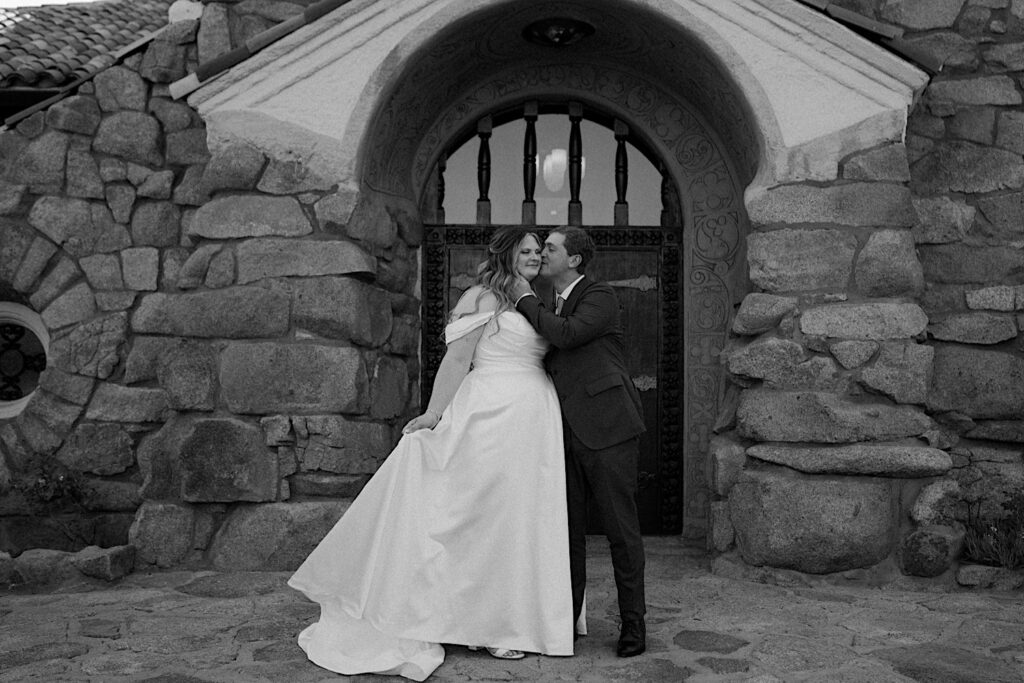 A black and white image of a groom kissing his bride on the cheek outside of their California wedding venue Mt Woodson Castle.  The bride holds her dress like a princess.