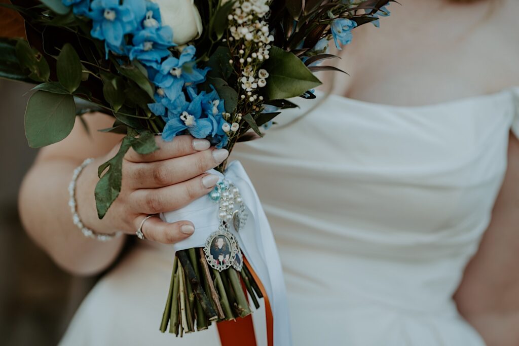 A bridal bouquet with blue and white flowers wrapped in white and orange ribbon.  She has memorial charms pinned to the bouquet with photos of loved ones who have passed away.