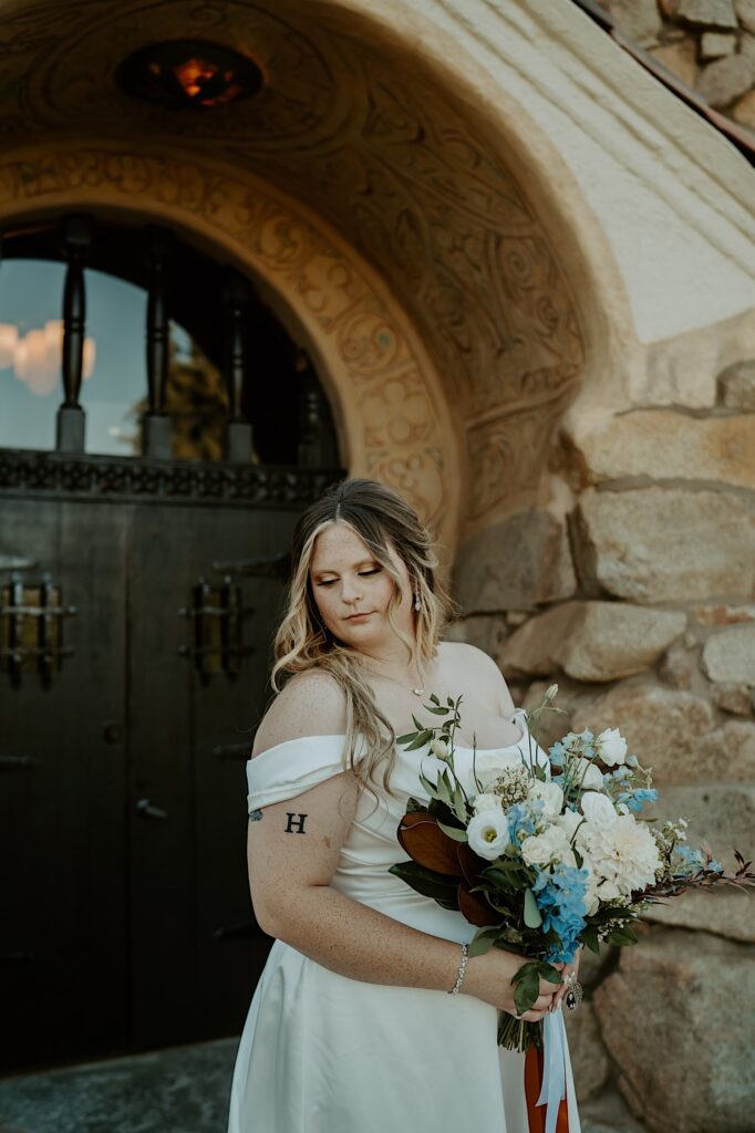 A bride stands with her wedding bouquet with blue and white flowers wrapped in orange and blue ribbon in front of her California Wedding Venue. 