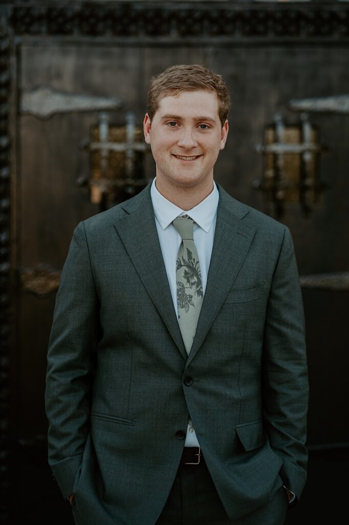 A groom with a charcoal grey wedding suit and a green tie with a floral pattern on it stands in front of his wedding venue in California.