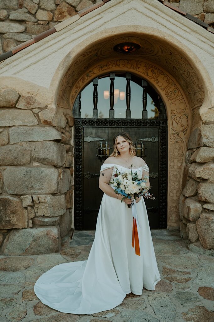 A bride stands with her wedding bouquet with blue and white flowers wrapped in orange and blue ribbon in front of her California Wedding Venue. 