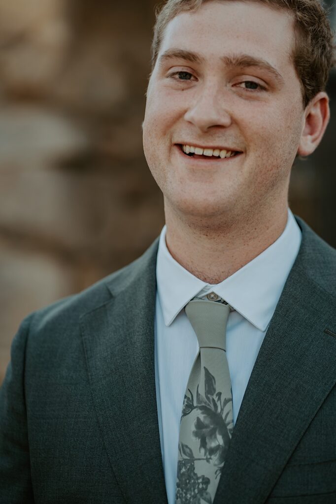 A groom with a charcoal grey wedding suit and a green tie with a floral pattern on it stands in front of his wedding venue in California.