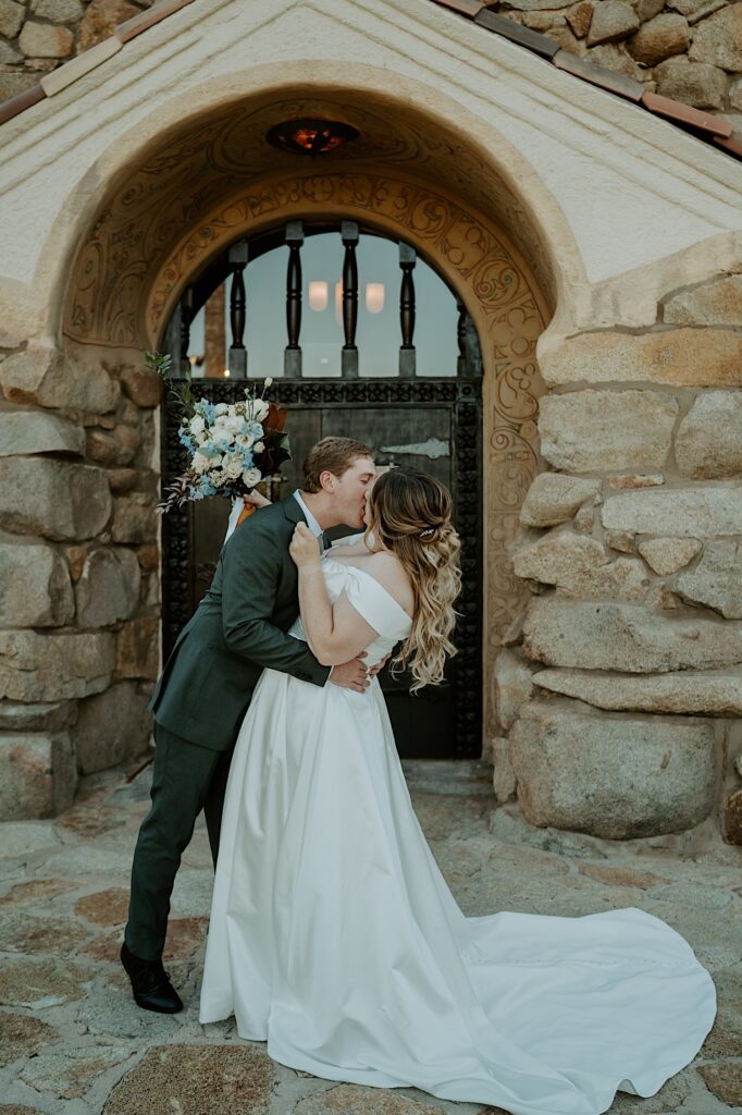 A bride and groom kiss outside of Mt Woodson Castle in California.  The bride has her hair in a half up half down hairstyle, and is holding a bridal bouquet with blue and white flowers.