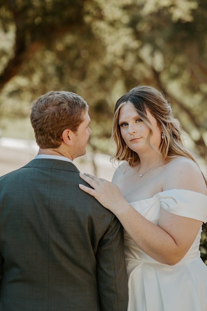 A bride and groom stand together, the groom is wearing a charcoal grey suit and he has his back to the camera while the bride is wearing a princess style wedding dress with her hair in a half up half down hairstyle.  She is looking at the camera.