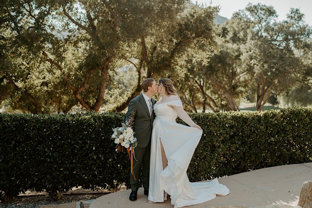 A bride and groom stand and kiss outside their California wedding venue.  The bride is wear a strapless dress with draping sleeves and the groom is holding her blue and white bouquet.