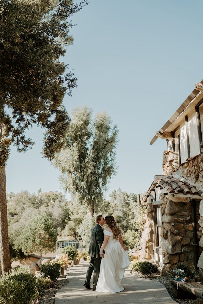 A bride and groom walk and hold hands and kiss outside of their California wedding venue Mt Woodson Castle. 