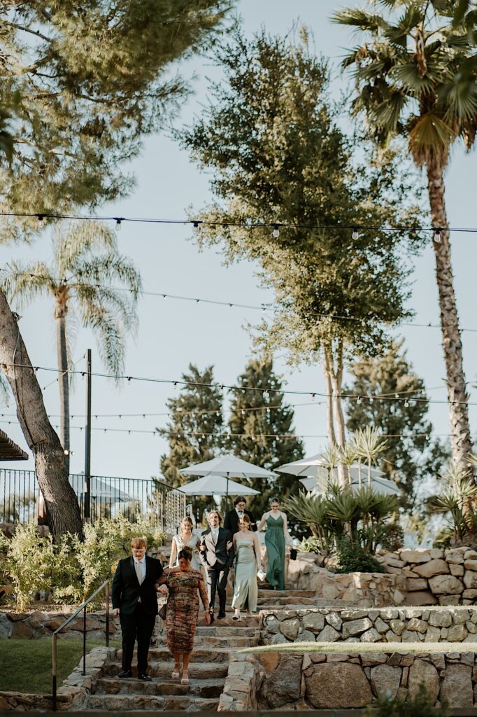 Guests walk into the outdoor wedding ceremony space at Mt Woodson Castle in California.  They walk down a set of stone steps in the California sun. 